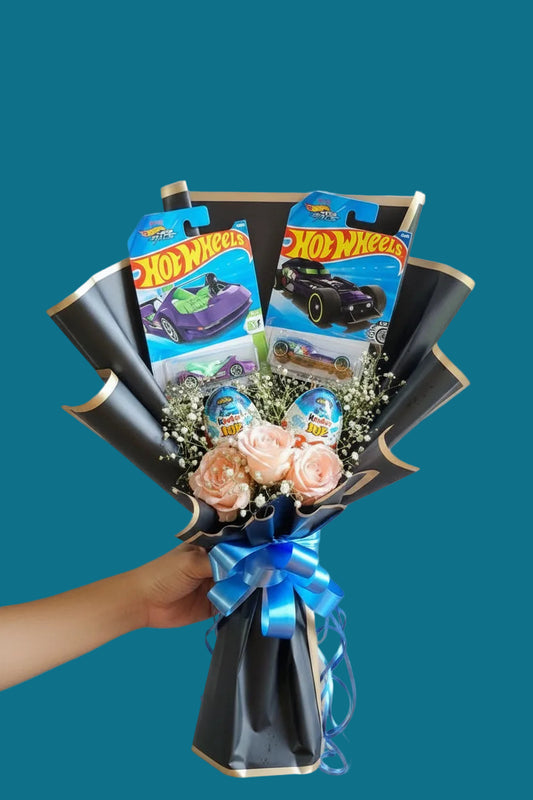 A Hot Wheels-themed gift bouquet held by a hand, featuring two Hot Wheels toy car packages, two Kinder joy, pink roses, baby's breath, and black paper petals tied with a blue ribbon, set against a purple curtain background.